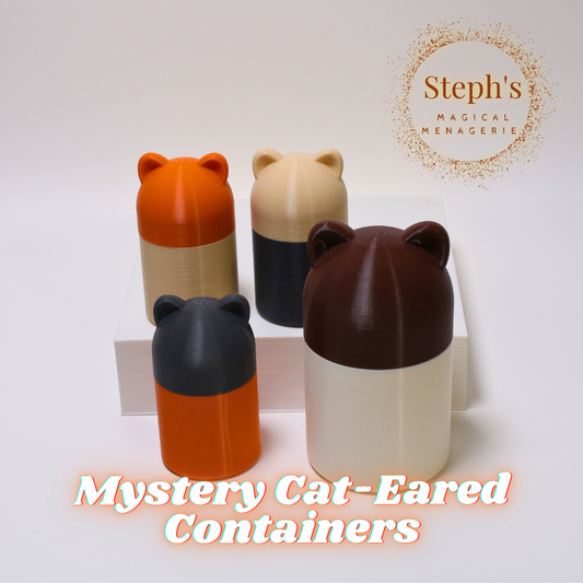 Four cylindrical containers with cat-ear-shaped lids, displayed on a white surface. Each container features a distinct color pairing: orange and beige, beige and dark gray, dark brown and beige, and dark gray and orange. Two containers are positioned on a raised platform, and one larger one sits on the lower surface next to another small container. The text 'Steph's Magical Menagerie' is located in the top right corner, with 'Mystery Cat-Eared Containers' written at the bottom.
